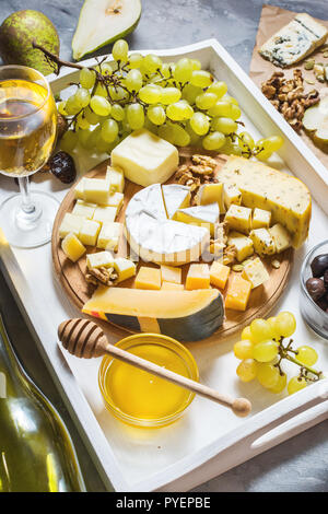 Different types of cheese on wooden board, olive, fruits, almond and wine glasses on white tray on concrete table Stock Photo