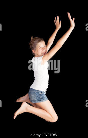 Young girl jumping in the air for fun and joy with arms stretched and legs bent isolated on black background wearing white shirt and denim shorts Stock Photo