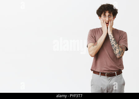 Gosh that is shocking. Portrait of stunned good-looking young urban male in shook with tattooed arm and moustache in glasses holding hands on face expressing empathy and worry over gray wall Stock Photo