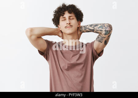 Guy feeling relieved stretching after sitting too long at work holding hands behind neck closing eyes lying back and enjoying evening being chill and calm having break over gray background Stock Photo