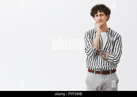 Man getting serious when talking about business. Portrait of determined focused and smart creative male with moustache tattoos and curly hair frowning poking cheek while thinking making decision Stock Photo