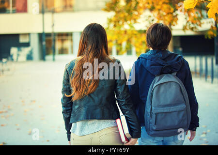 Rear view of two women students going to school or college carrying backpack and holding books in hand. Autumn outdoors friends walking educational co