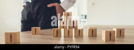 Retro vintage image of businessman standing next to an office desk with wooden cubes with people icons placed in  a pyramid. Stock Photo