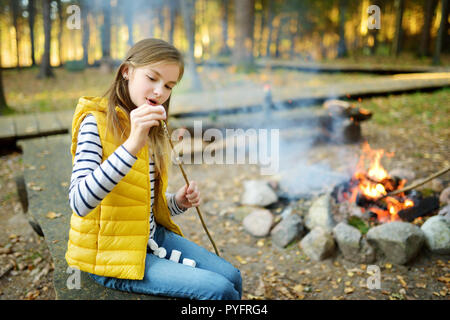 Cute preteen girl roasting marshmallows on stick at bonfire. Child having fun at camp fire. Camping with children in fall forest. Family leisure with  Stock Photo