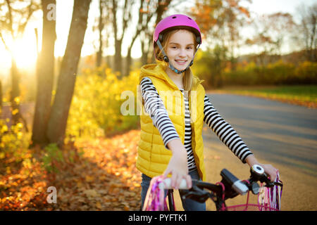 Cute little girl riding a bike in a city park on sunny autumn day. Active family leisure with kids. Child wearing safety hemet while riding a bicycle. Stock Photo