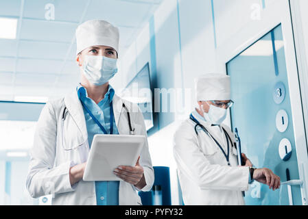 low angle view of female doctor in medical mask using digital tablet while her male colleague standing behind with clipboard in hospital corridor Stock Photo