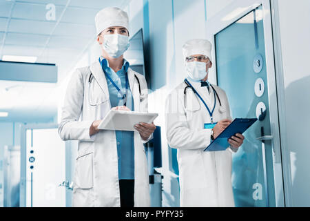 low angle view of female doctor in medical mask using digital tablet while her male colleague standing behind with clipboard in hospital corridor Stock Photo