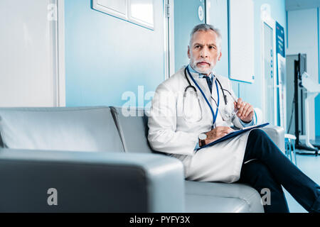 middle aged male doctor with clipboard looking away and sitting on couch in hospital corridor Stock Photo