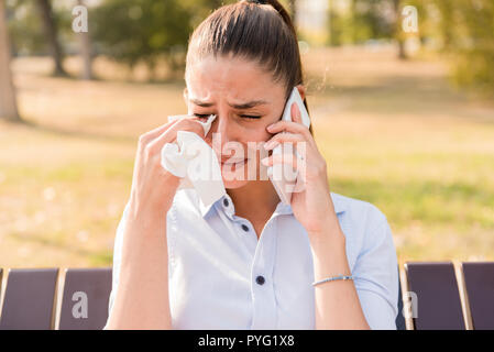 Sad young woman cries while talking on the mobile phone in the park on bench Stock Photo