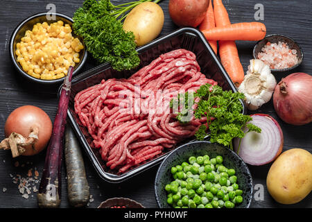 Raw minced meat in container surrounded by ingredients for shepherds pie , green peas, yellow corn, carrot, onion and seasonings, on black background, Stock Photo