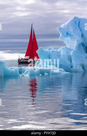 Beautiful red sailboat in the arctic next to a massive iceberg showing the scale. Ilulissat, Disko Bay, Greenland. Stock Photo