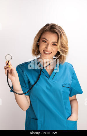 Female smiling doctor displaying a stethoscope to the camera and hand in the pocket. Cheerful medic doing his job Stock Photo