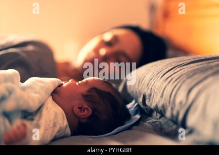 A baby asleep on bed with his mum in the background. Stock Photo