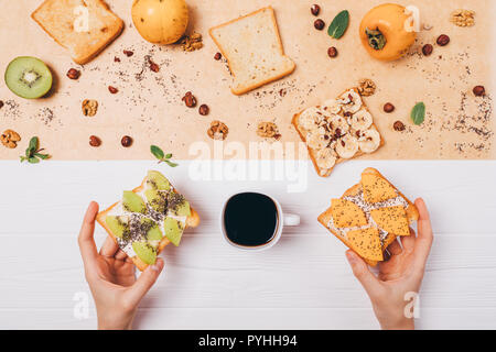 Woman's hands holding two toast with fruit next to cup of black coffee, top view. Food composition healthy sandwiches with kiwi, banana and persimmon  Stock Photo