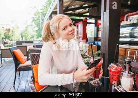 young girl with freckles texting message on her smartphone while drinking coctail with berries and mint in the outdoor restaurant