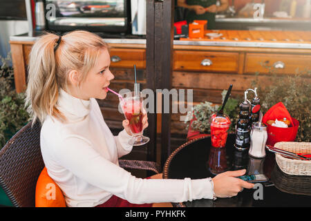 young girl with freckles texting message on her smartphone while drinking coctail with berries and mint in the outdoor restaurant Stock Photo