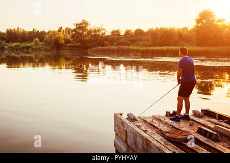 Young man fishing on river standing on bridge at sunset. Happy fiserman enjoying hobby in summer Stock Photo