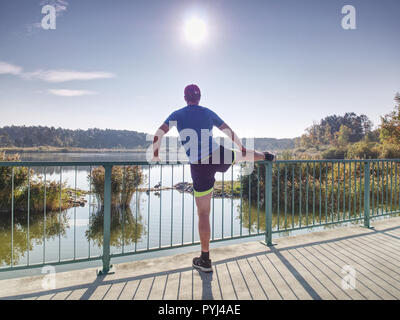 Man stretching legs before going  run outside within sunny morning. Warming up muscles. Outdoor exercising on lake bridge.