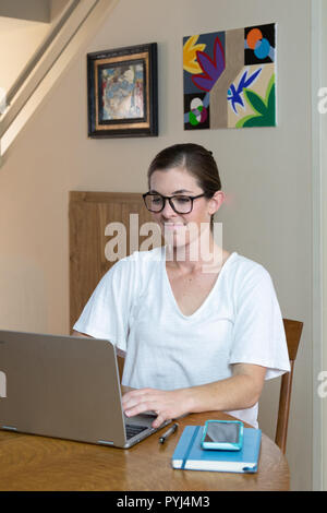 Attractive young woman working on computer at dining room table with her artwork/painting on the wall behind and technological devices. Stock Photo
