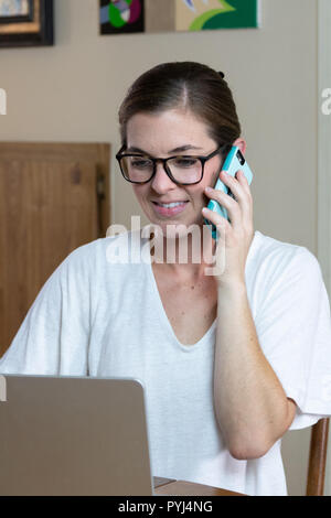 Close up image of young woman working on computer at dining room table talking on cell phone with teeth showing and pleasant look on her face. Stock Photo