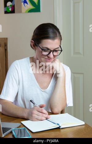 Young woman artist/painter reading journal with cell phone and laptop resting chin on hand close up sitting at dining room table. Stock Photo