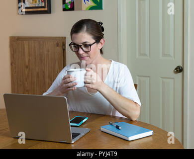 Young woman artist and painter working looking down at laptop holding a mug to take a drink with communication devices on table in front of her. Stock Photo