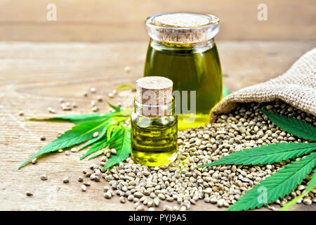 Hemp oil in two glass jars with grain in the sack, leaves and stalks of cannabis on the background of old wooden boards Stock Photo