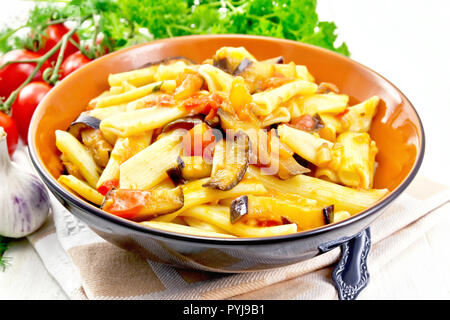 Penne pasta with eggplant and tomatoes in a bowl on towel, fork, garlic and parsley on a wooden plank background Stock Photo