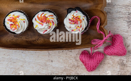 3 chocolate cupcakes with vanilla frosting and sprinkles on a wooden tray with pink knitted hearts on a light wood background Stock Photo