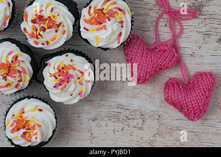 chocolate cupcakes with white frosting and sprinkles, knitted pink  hearts on a light wood background Stock Photo