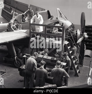 1960s, historical, male engineer in whitecoat supervising a number of young aviaiton apprentices as they work on the cockpit of a single-propellered aircraft. Stock Photo