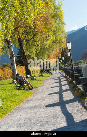 People rest on benches in the sunshine among autumn colored trees on the lakefront promenade at Lake Schliersee, Bavaria, Germany Stock Photo