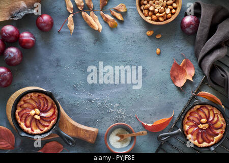 Cooking background with red plums, plum crumble cakes, towel, caramelized peanuts, autumn leaves. Top view with copy-space on gray textured background Stock Photo