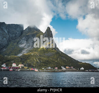Famous tourist attraction of Reine in Lofoten with view towards Reinebringen, Norway with red rorbu houses, clouds, rainy day with bridge and grass an Stock Photo