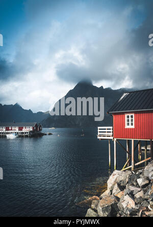 Rainbow ofer red houses rorbuer of Reine in Lofoten, Norway with red rorbu houses, clouds, rainy blue sky and sunny. Bridges and mountains in the back Stock Photo