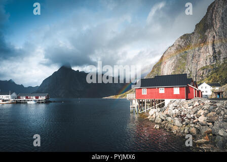 Rainbow ofer red houses rorbuer of Reine in Lofoten, Norway with red rorbu houses, clouds, rainy blue sky and sunny. Bridges and mountains in the back Stock Photo