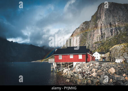 Rainbow ofer red houses rorbuer of Reine in Lofoten, Norway with red rorbu houses, clouds, rainy blue sky and sunny. Bridges and mountains in the back Stock Photo