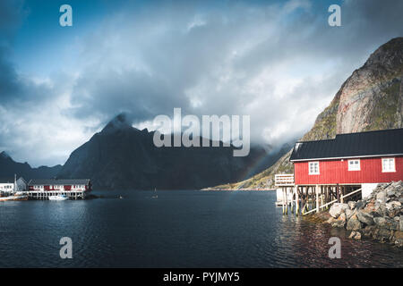 Rainbow ofer red houses rorbuer of Reine in Lofoten, Norway with red rorbu houses, clouds, rainy blue sky and sunny. Bridges and mountains in the back Stock Photo