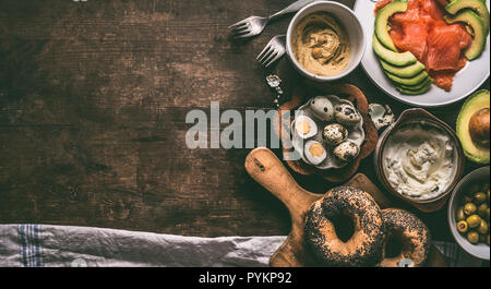 Home breakfast preparation with bagel bread, salmon, avocado , fresh cheese, hummus and cooked quail eggs on dark rustic wooden background, top view.  Stock Photo
