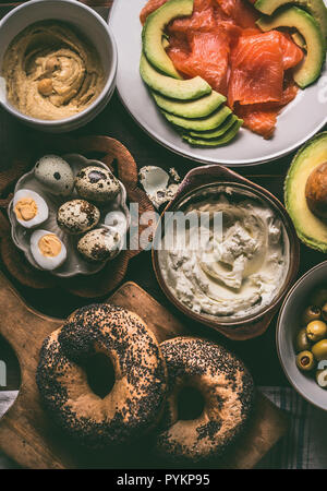 Breakfast preparation with bagel bread, salmon, avocado , fresh cheese, hummus and cooked quail eggs on dark rustic wooden background, top view. Stock Photo