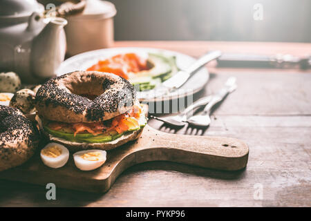 Homemade bagel sandwich topped with salmon, avocado , fresh cheese and cooked quail eggs on dark rustic wooden background. Breakfast or snack preparat Stock Photo