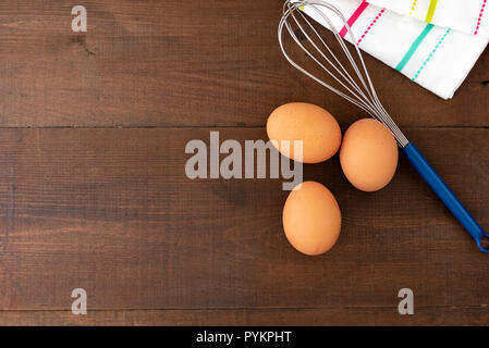 Three eggs a a wire steel whisk, on a wooden background, with copy space. Stock Photo
