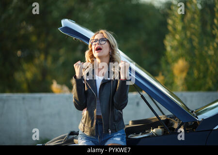 Image of frustrated woman in glasses near broken car with open hood Stock Photo