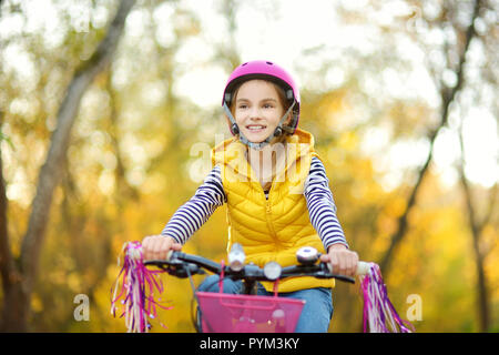 Cute little girl riding a bike in a city park on sunny autumn day. Active family leisure with kids. Child wearing safety hemet while riding a bicycle. Stock Photo