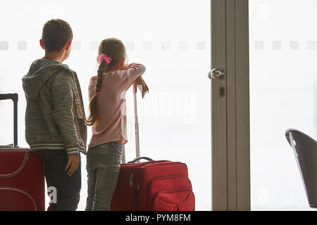 Two children in the airport terminal look curiously through a window on the runway Stock Photo