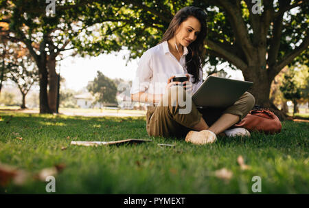 Beautiful woman sitting on grass lawn at park holding a coffee and looking at laptop computer. Young businesswoman in casual clothes sitting at park w Stock Photo