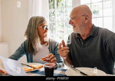 Senior man and woman talking to each other while eating breakfast at home. Woman holding a paper discussing with her husband at the breakfast table. Stock Photo