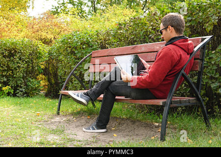 A young man in casual clothes in a warm jacket working outdoors in the autumn Park, read a book on ultrabook. teenager searching media content togethe Stock Photo