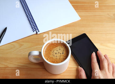 Female's Hand Holding Cellphone beside a Cup of Hot Coffee with a Notebook and Pen on the Working Desk Stock Photo