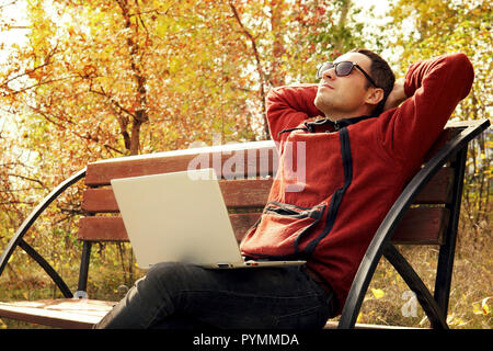 Happy mature man using laptop while sitting on a bench in a city Park. Young it specialist take a break from work. Remote work outdoors. The student i Stock Photo